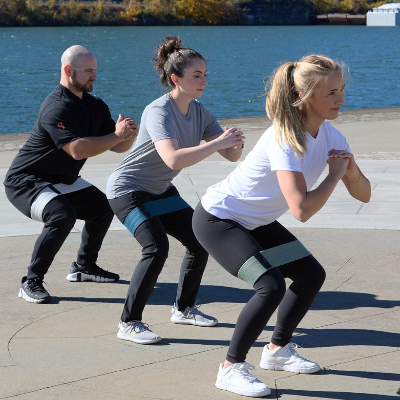 Three people performing a workout routine by a waterfront using some resistance bands.